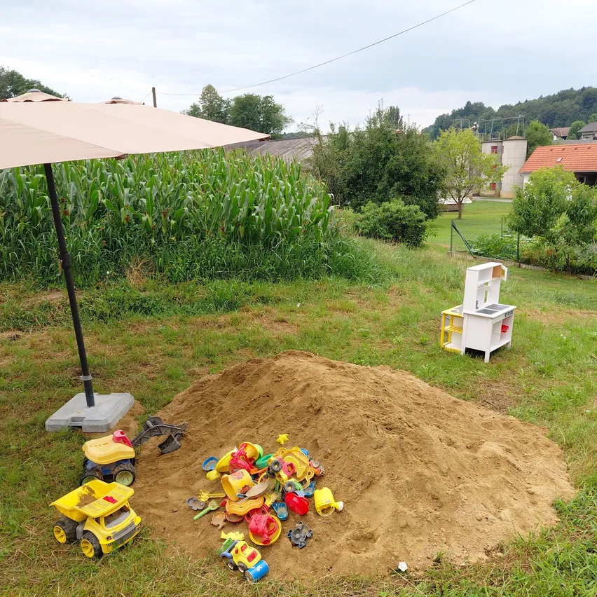 Ein Garten mit einem Sandkasten voller Spielzeug, einem weißen Spielhaus und einem Sonnenschirm in einem ländlichen Gebiet.