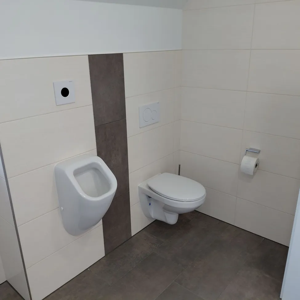 Ein sauberes, minimalistisches Badezimmer mit einem weißen Urinal, einem an der Wand montierten WC und einer weißen gefliesten Wand mit grauem Akzent. Ein Toilettenpapierhalter ist an der Wand montiert.