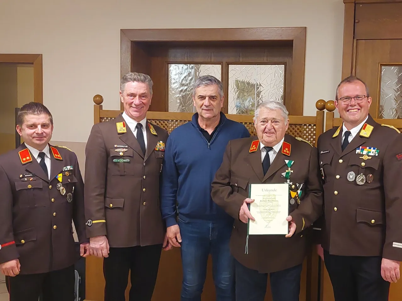 Fünf Männer stehen vor einer Holztür mit Glasscheiben. Der Mann in der Mitte hält ein Zertifikat und trägt eine Medaille. Die anderen Männer sind in Uniformen.
