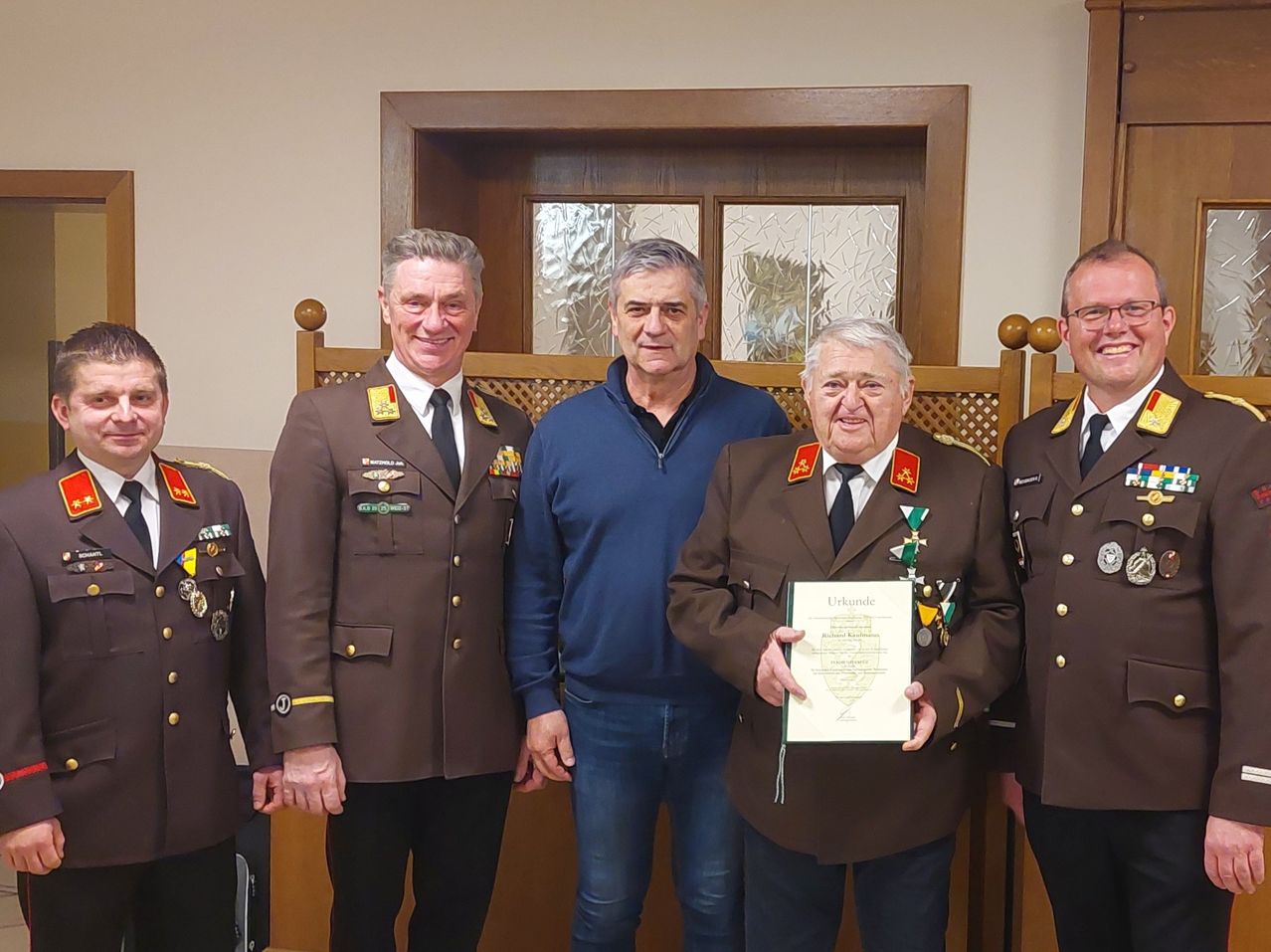 Fünf Männer stehen vor einer Holztür mit Glasscheiben. Der Mann in der Mitte hält ein Zertifikat und trägt eine Medaille. Die anderen Männer sind in Uniformen.