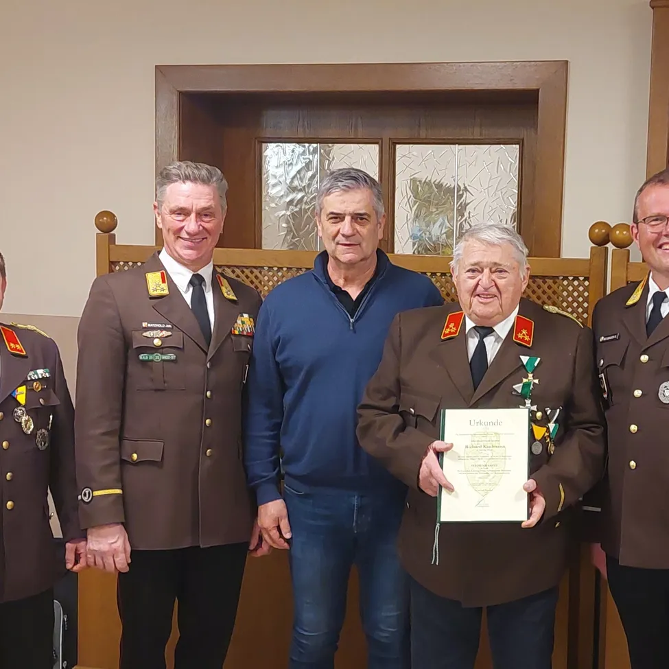 Fünf Männer stehen vor einer Holztür mit Glasscheiben. Der Mann in der Mitte hält ein Zertifikat und trägt eine Medaille. Die anderen Männer sind in Uniformen.