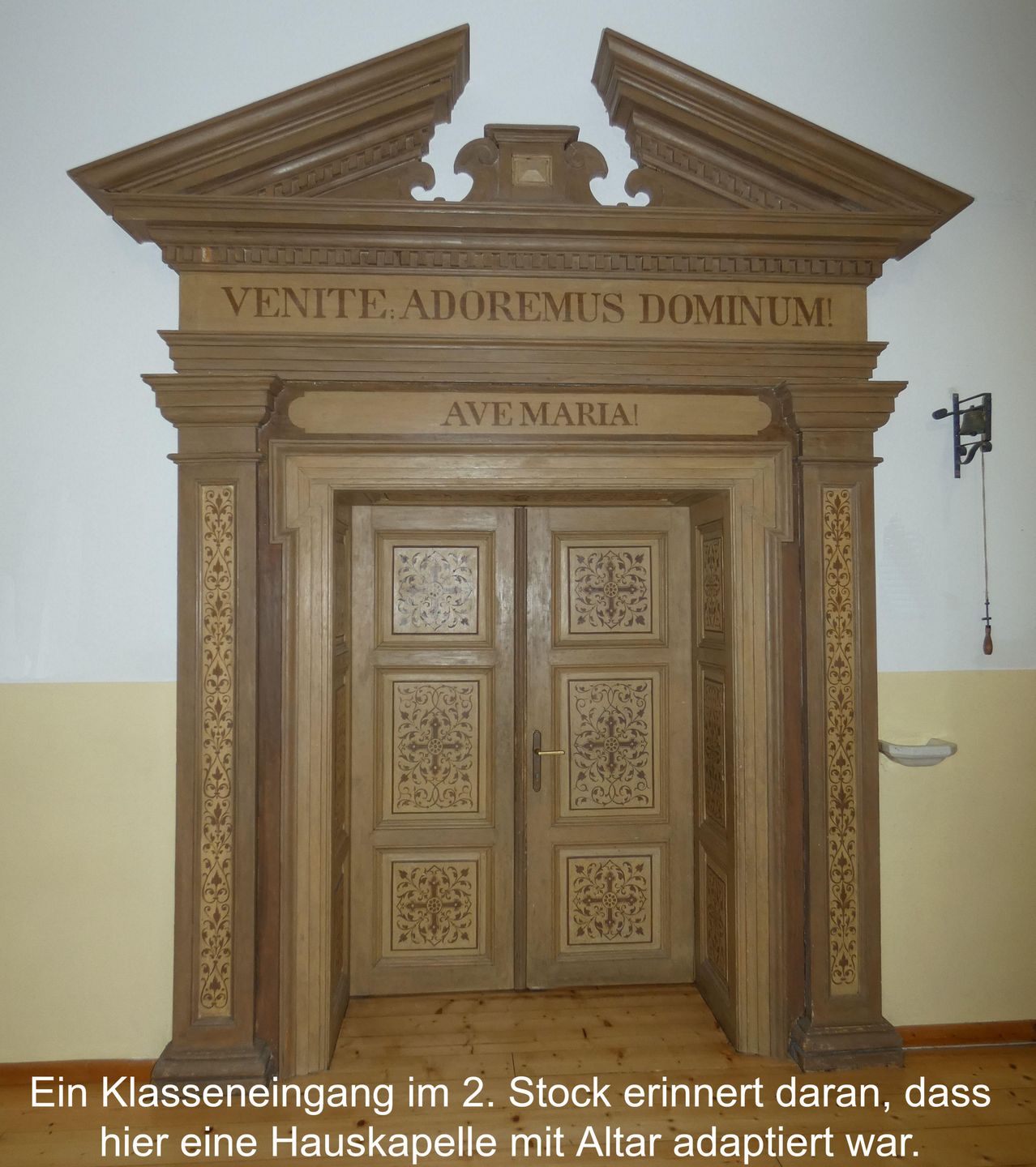 Eine Holztür mit einem kunstvollen Rahmen und lateinischen Inschriften darüber. Die Tür hat aufwendige Schnitzereien und einen Messinggriff. Sie führt zu einem Holzboden und wird von einem Bilduntertext begleitet.