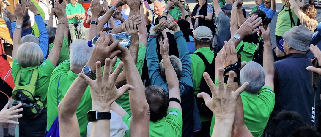 Eine Menschenmenge in grünen Shirts hebt die Hände, möglicherweise zum Jubeln. Eine Frau in der Mitte mit Brille und Halskette. Ein roter Lieferwagen ist im Hintergrund geparkt.