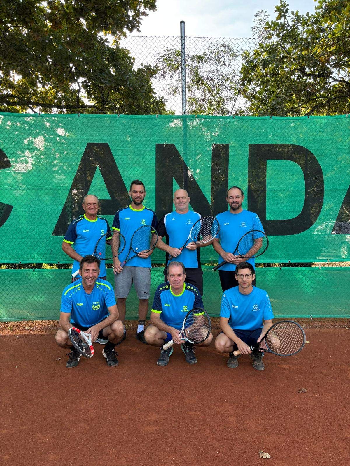 Eine Gruppe von Männern in blauen Trikots posiert für ein Foto auf einem Tennisplatz, mit einem grünen Banner, das 'ANDA' hinter ihnen steht.