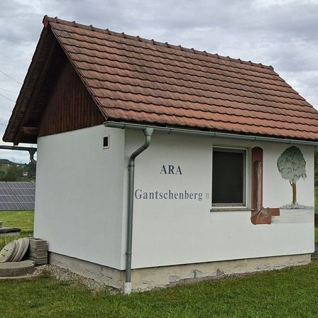 Ein kleines weißes Gebäude mit rotem Dach befindet sich auf einem Rasenfeld. Das Gebäude hat einen Baum an der Seite und Solarmodule im Hintergrund.