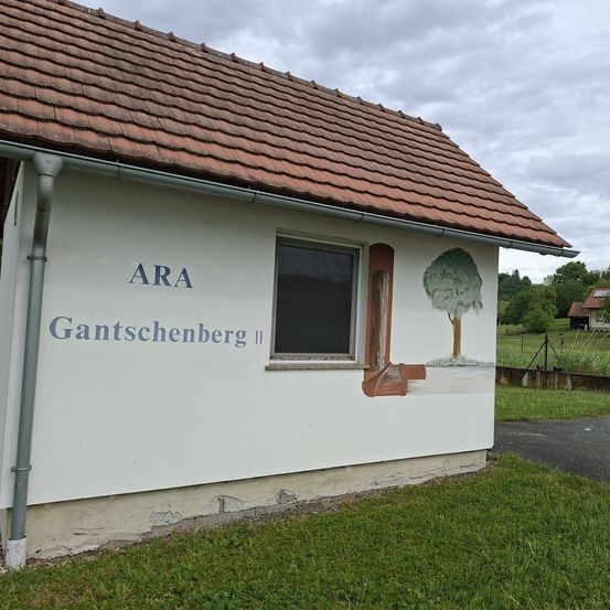 Ein Gebäude mit einem braunen Dach und weißen Wänden. Die Worte ARA Gantschenberg II sind darauf gemalt. Es gibt ein Fenster und eine Baummalerei an der Wand.