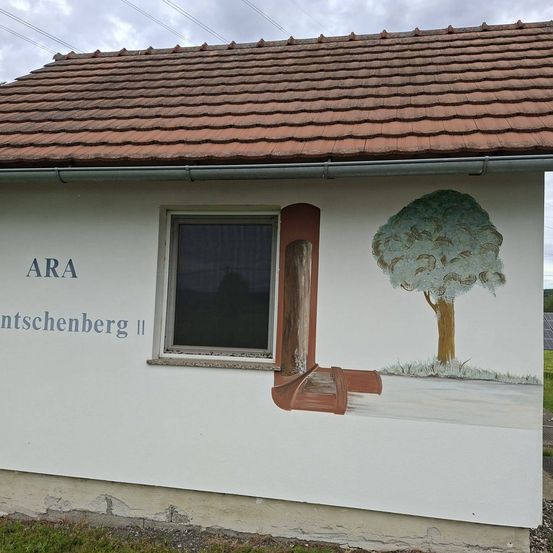 Ein weißes Gebäude mit einem braunen Dach und einem Fenster. Die Wand zeigt ein Gemälde eines Baumes und einer offenen Tür. Die Worte 'ARA' und 'ntschengberg II' sind an der Wand geschrieben.