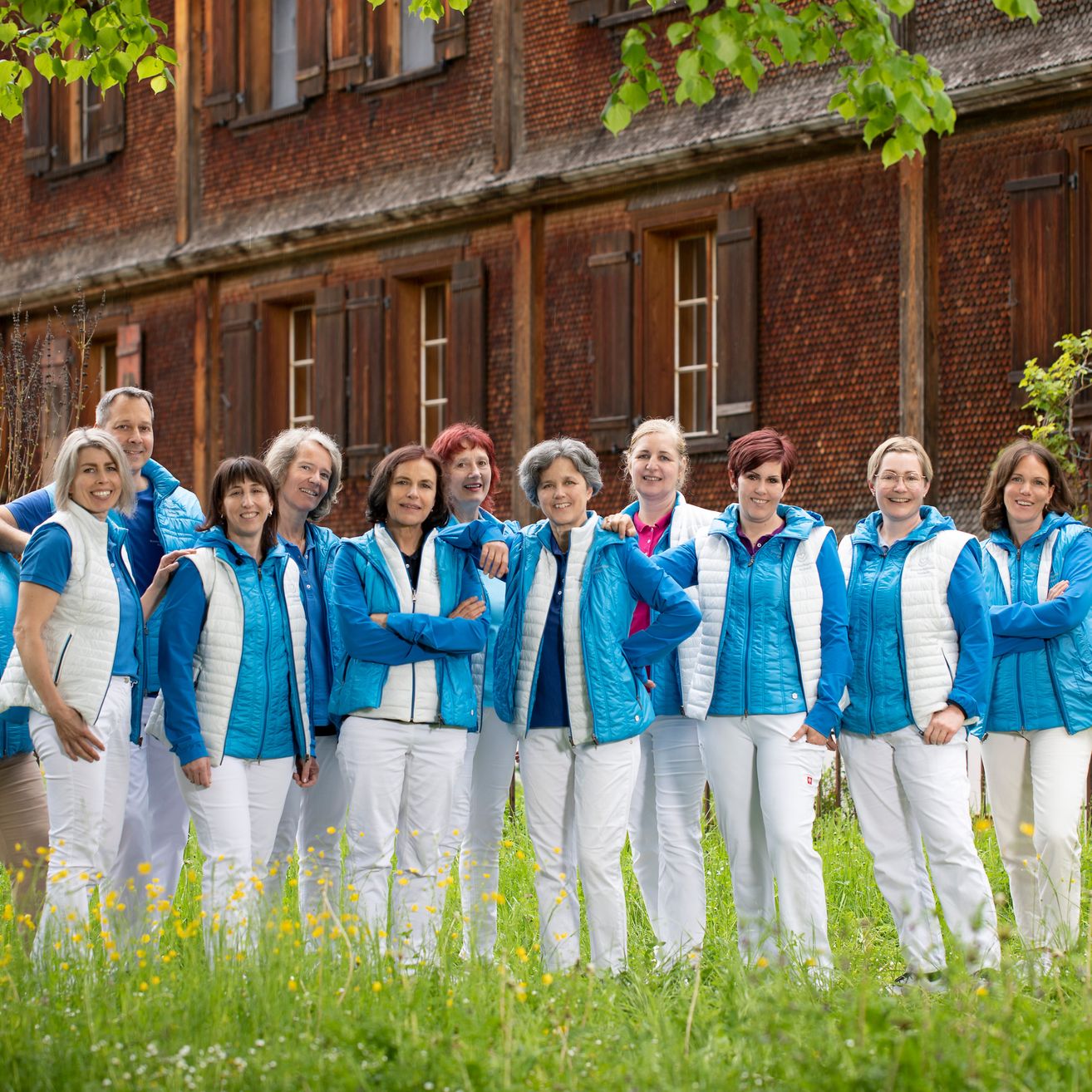 A group of women in matching blue and white jackets and pants stand together in a field, smiling for a photo in front of a brick building.