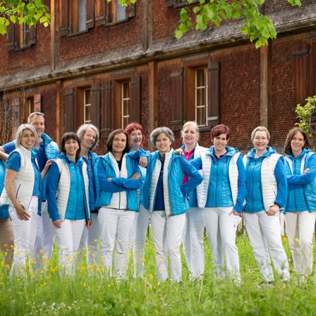 Eine Gruppe von Frauen in passenden blauen und weißen Jacken und Hosen steht zusammen in einem Feld und lächelt für ein Foto vor einem Backsteingebäude.