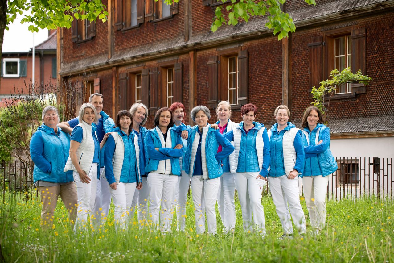 Eine Gruppe von Frauen in blauen und weißen Jacken und weißen Hosen steht auf einem Rasenfeld vor einem Backsteingebäude.
