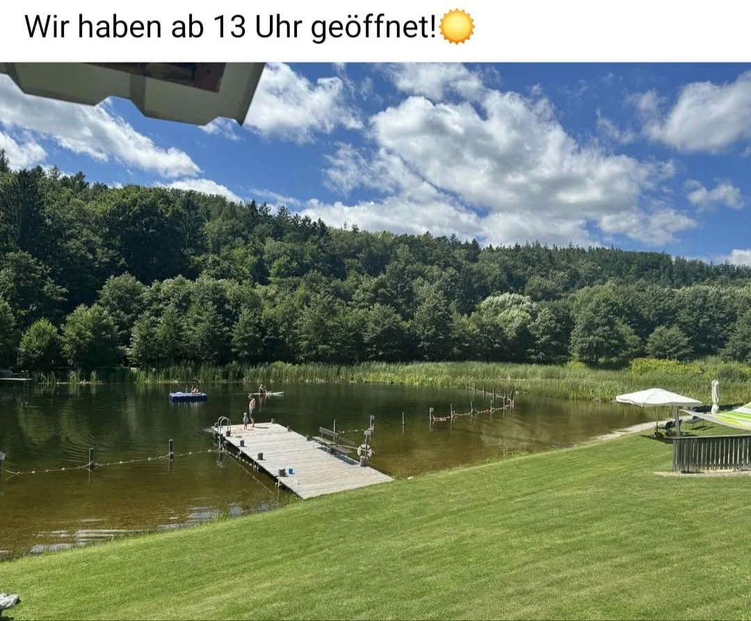Ein See mit einem Steg und einer Person, die in der Nähe des Wassers steht. Ein kleines Boot schwimmt auf dem See.