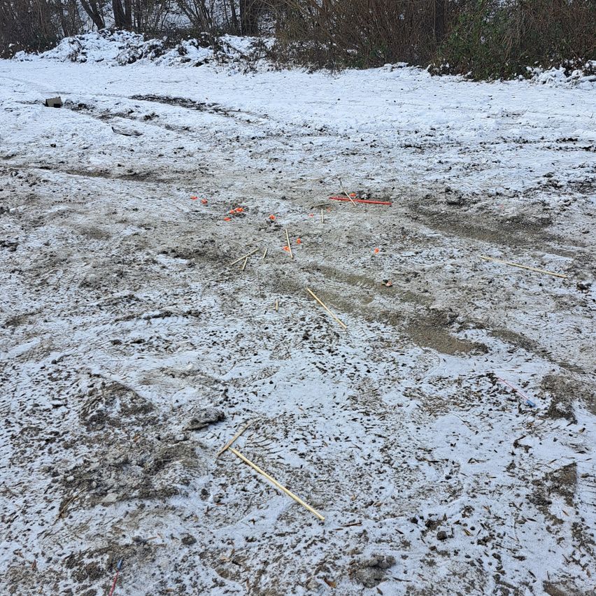 Eine verschneite Landschaft mit einer Reifenspur in der Mitte, umgeben von orangefarbenen Markierungen und Stöcken, möglicherweise als Suchbereich markiert.