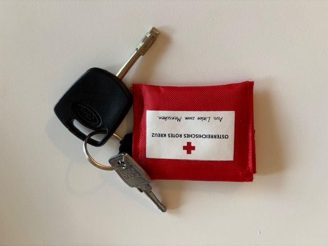 A red first aid kit with a white label is attached to a black car key. The label reads, 'Kreuz Roter Schweizer Kreuz'. The key has a silver ring and a keyhole.