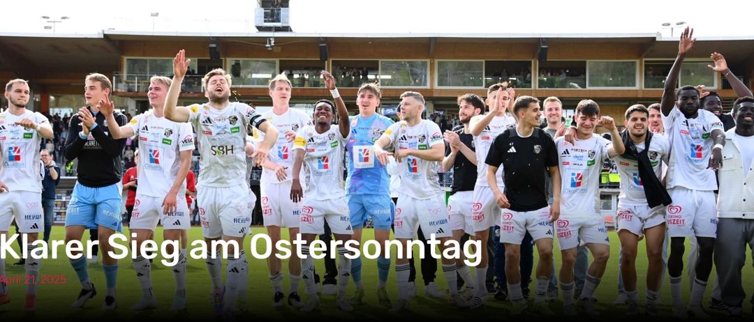 Gruppe männlicher Fußballspieler feiert einen Sieg auf einem Rasenfeld. Sie tragen weiße Trikots mit verschiedenen Team-Logos. Der Text lautet 'm Ostersonntag'. Hinter ihnen ist ein verschwommener Hintergrund eines Gebäudes mit Glasfenstern und Menschen.