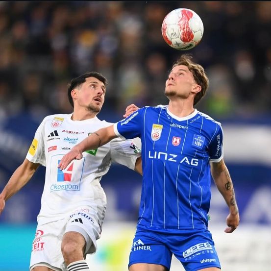 Zwei Fußballspieler in Trikots versuchen, den Ball zu köpfen. Der Spieler in Blau schaut nach oben, während der Spieler in Weiß nach unten schaut.
