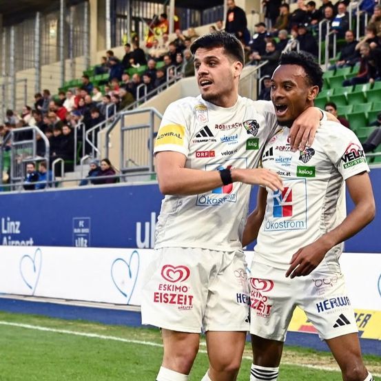 Zwei Fußballspieler feiern ein Tor auf dem Spielfeld. Sie tragen weiße Trikots mit verschiedenen Werbungen. Hinter ihnen ist das Stadion voll mit Zuschauern, und die Tribünen sind grün und blau bemalt.