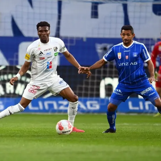 Zwei Fußballspieler spielen ein Spiel. Der Spieler in Weiß versucht, den Ball zu schießen, während der Spieler in Blau versucht, ihn zu blockieren. Das Spielfeld hat eine grüne Rasenfläche und einen unscharfen Hintergrund.