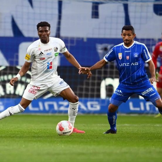 Zwei Fußballspieler spielen ein Spiel. Der Spieler in Weiß versucht, den Ball zu schießen, während der Spieler in Blau versucht, ihn zu blockieren. Das Spielfeld hat eine grüne Rasenfläche und einen unscharfen Hintergrund.