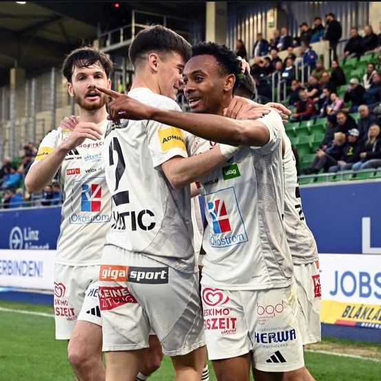 Drei männliche Fußballspieler in weißen Trikots mit den Nummern 4, 6 und 10 feiern auf dem Spielfeld. Der Spieler mit der Nummer 10 hat seinen Arm um den Spieler mit der Nummer 6. Sie stehen vor einer Menge in den Rängen.