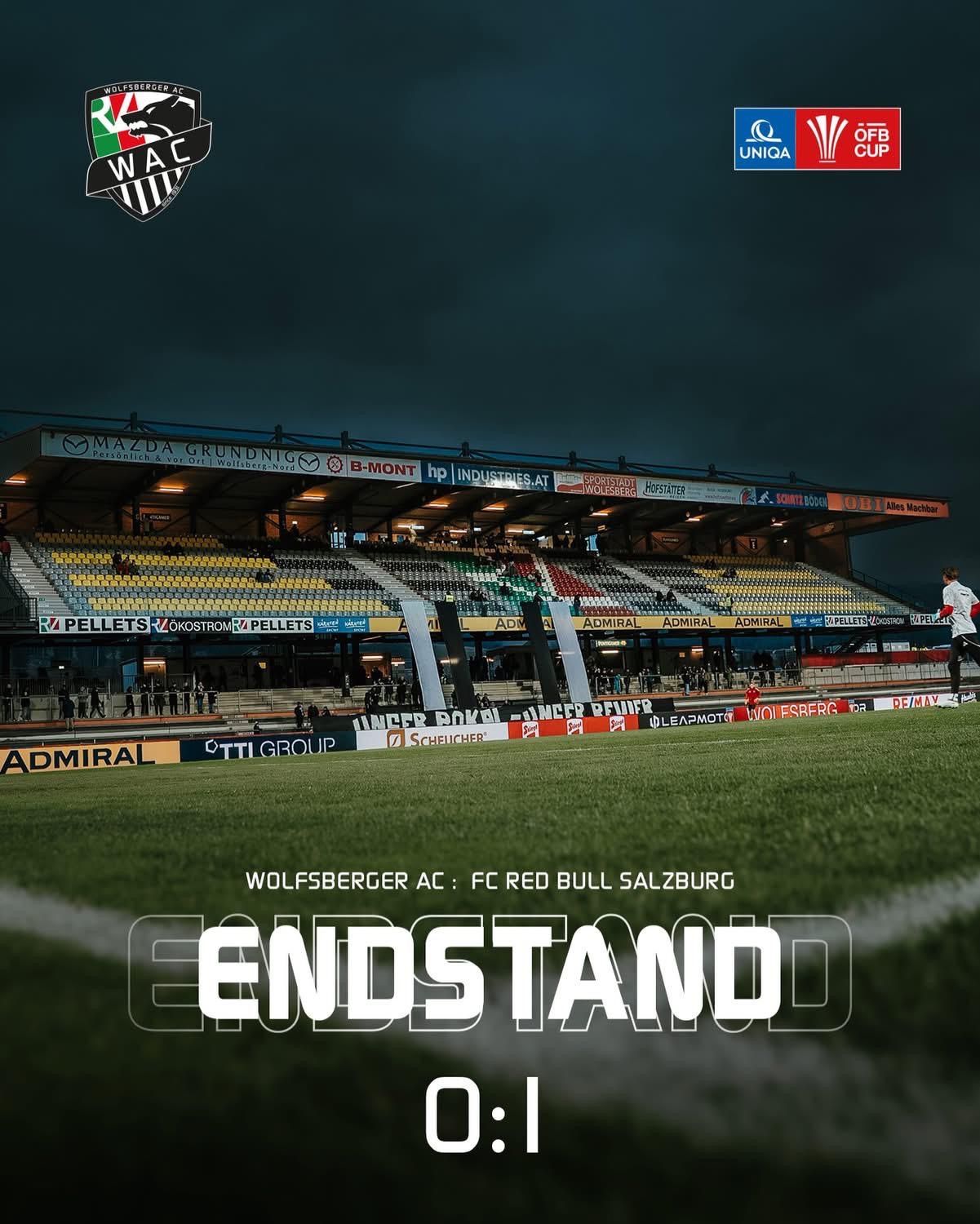 Ein Fußballfeld mit einer Anzeigetafel, die ein Spiel zwischen Wolfsberger AC und FC Red Bull Salzburg anzeigt. Das Feld ist leer, und ein Spieler läuft im Hintergrund.