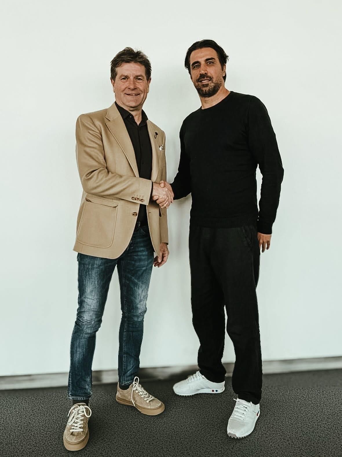 Two men standing and shaking hands, one wearing a beige jacket and the other in a black long-sleeve shirt and black pants.