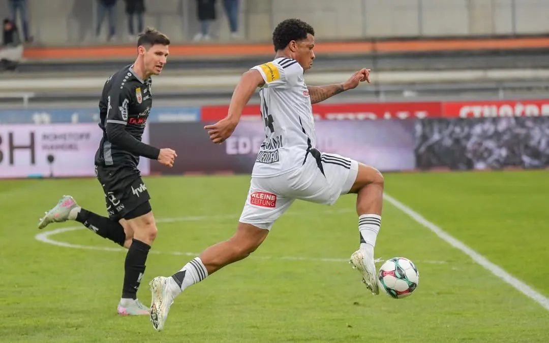 Zwei Fußballspieler in Aktion auf dem Feld. Ein Spieler springt und tritt den Ball, während der andere hinter ihm herläuft.