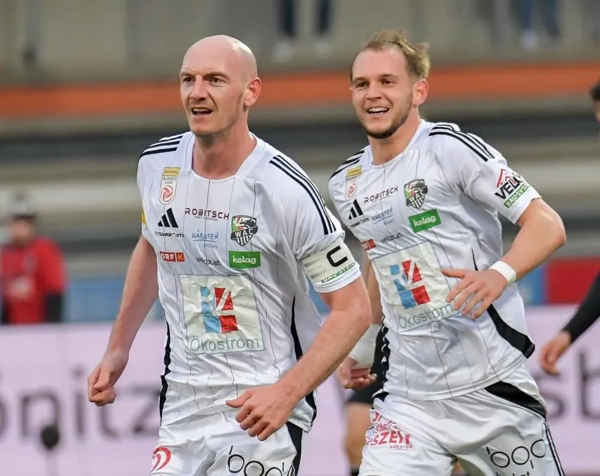 Zwei Fußballspieler in weißen Trikots feiern auf dem Feld, einer mit einem Trikot der Nummer 20 und der andere mit einem Trikot der Nummer 14.