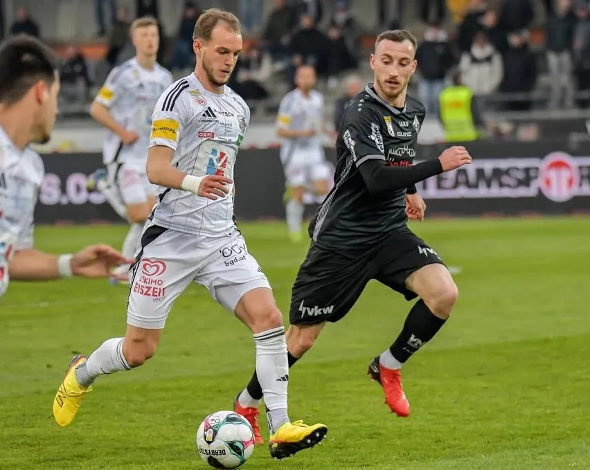 Zwei Fußballspieler in Aktion auf dem Feld, einer jagt dem anderen hinterher und hält den Ball.