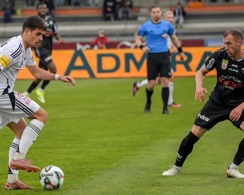 Ein Fußballspiel mit Spielern in schwarzen, weißen und blauen Uniformen. Der Ball liegt auf dem Boden und die Spieler rennen darauf zu. Im Hintergrund steht ein Banner mit der Aufschrift 'ADMR'.
