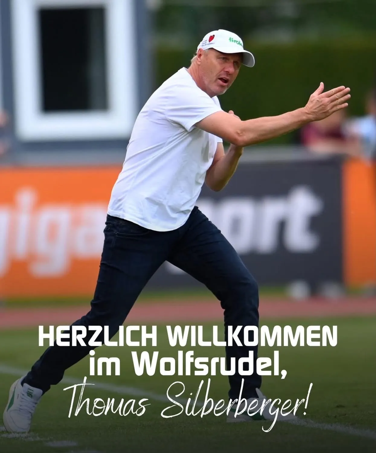 Ein Mann in einem weißen Shirt und einer Kappe steht auf einem Fußballfeld und gestikuliert mit den Armen. Hinter ihm steht ein orangefarbenes Banner mit Gigasport. Der Text auf dem Bild lautet Herzlich Willkommen im Wolfsrudel, Thomas Silberberger!