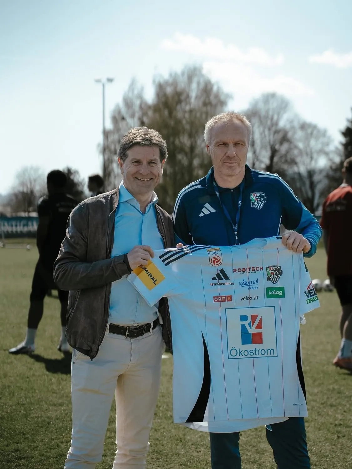 Zwei Männer stehen auf einem Feld, einer hält ein weißes Trikot mit mehreren Logos. Sie lächeln und posieren für ein Foto. Hinter ihnen sind verschwommene Personen auf dem Feld.