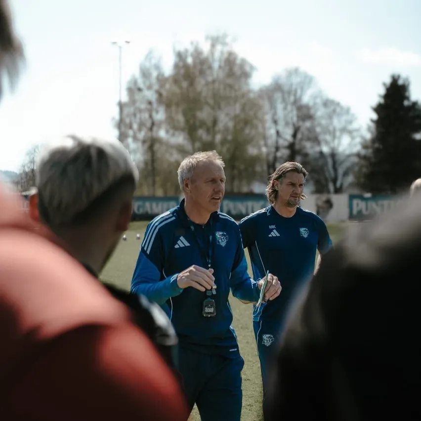 Zwei Männer in blauen Uniformen unterhalten sich auf einem Feld, umgeben von anderen Personen, Bäumen und einer entfernten Straßenlaterne.