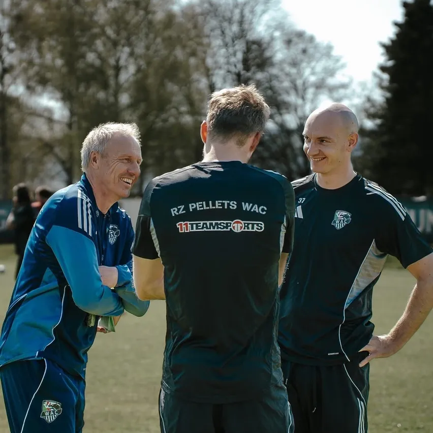 Drei Männer stehen draußen auf einem Feld, zwei tragen grüne und weiße Sportuniformen, einer trägt blau und weiß. Die beiden in Grün lächeln und reden. Dahinter sind Bäume und Menschen.