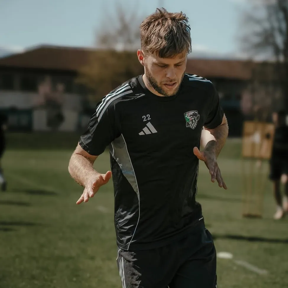 Ein Mann in einem schwarzen Adidas-Trikot mit der Nummer 22 läuft auf einem Feld. Er schaut nach links. Im Hintergrund befinden sich Bäume und ein Gebäude.