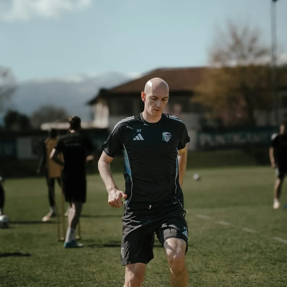 Ein Fußballspieler läuft über ein grünes Feld. Dahinter sind andere Spieler, und im Hintergrund steht ein Haus mit einem Baum.