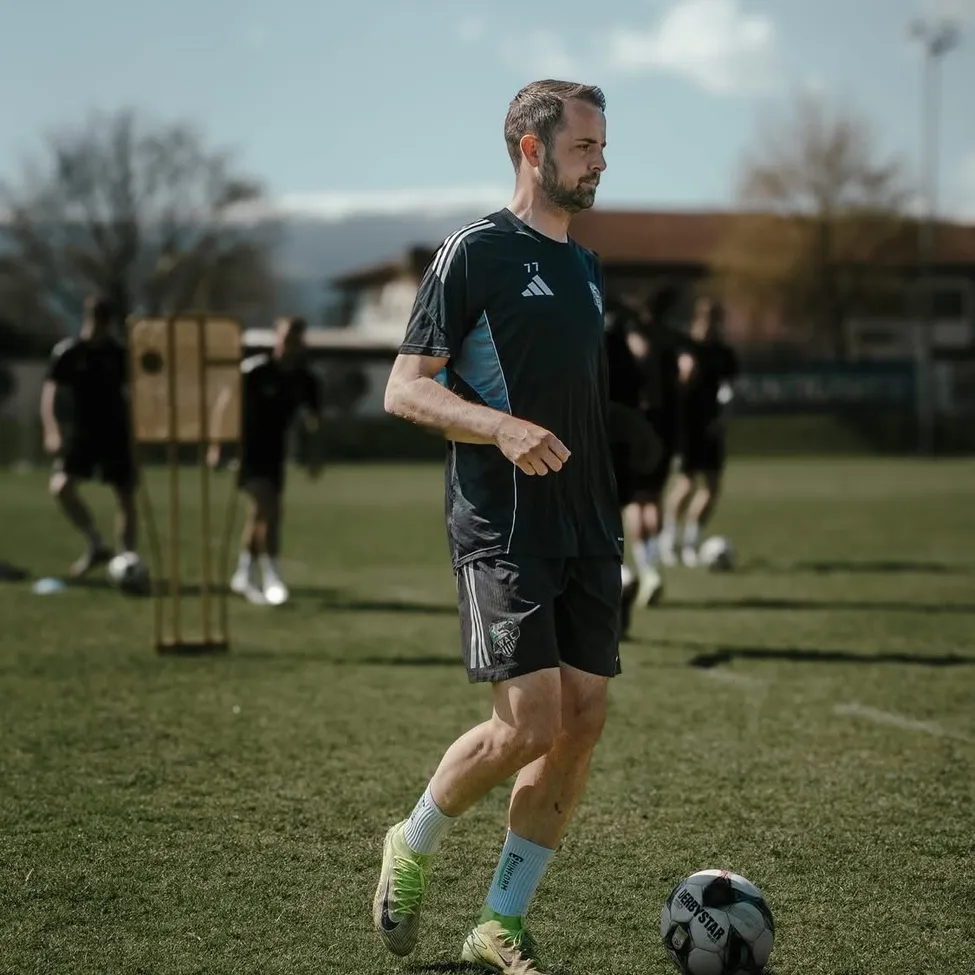 Ein Fußballspieler in schwarz-blauer Sportkleidung trainiert auf einem Feld, während andere Spieler im Hintergrund sind.