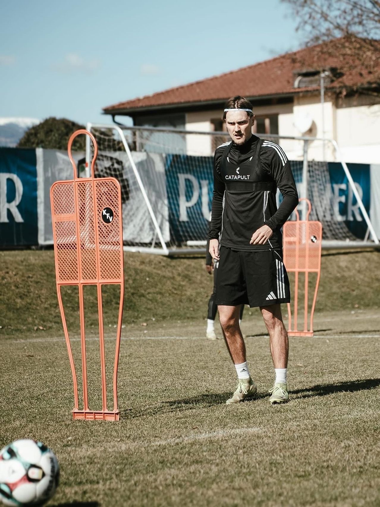 A soccer player is standing on a grassy field, wearing a black outfit. He is looking sideways, possibly towards the camera. Orange goalposts are positioned on the field. In the background, there is a building with a brown roof and a banner.