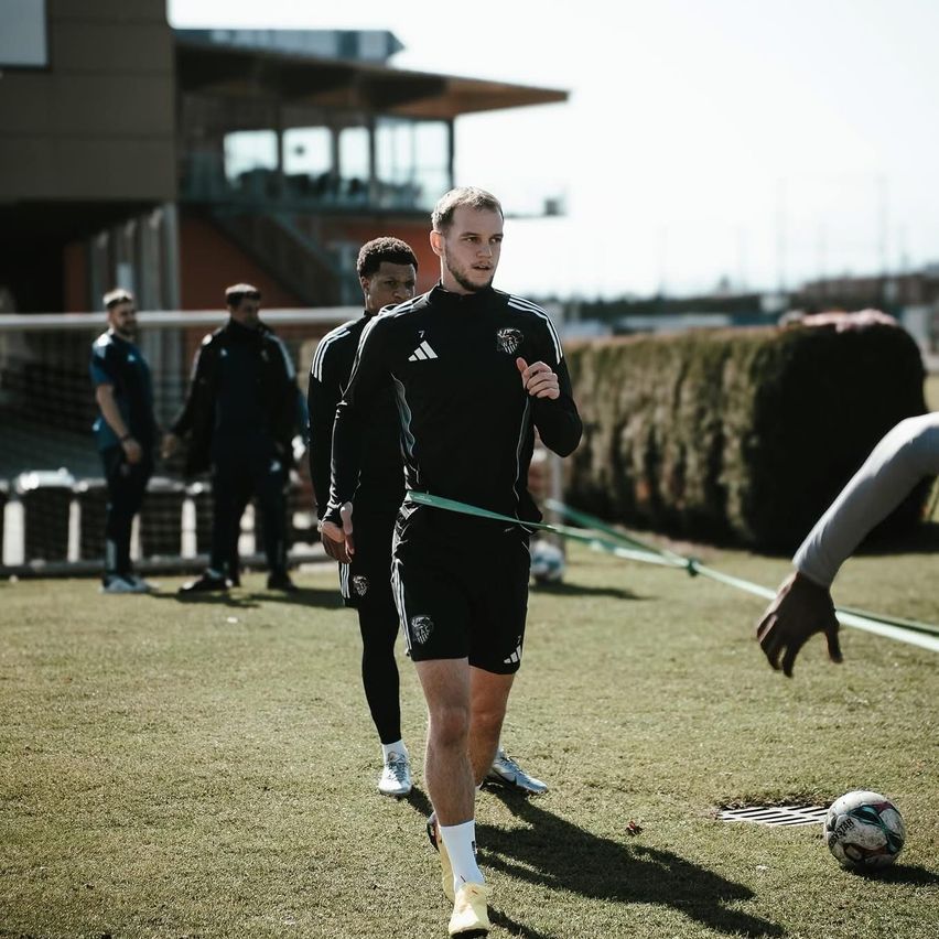 A soccer player in a black uniform is jogging on the field, while others observe. A soccer ball lies nearby.