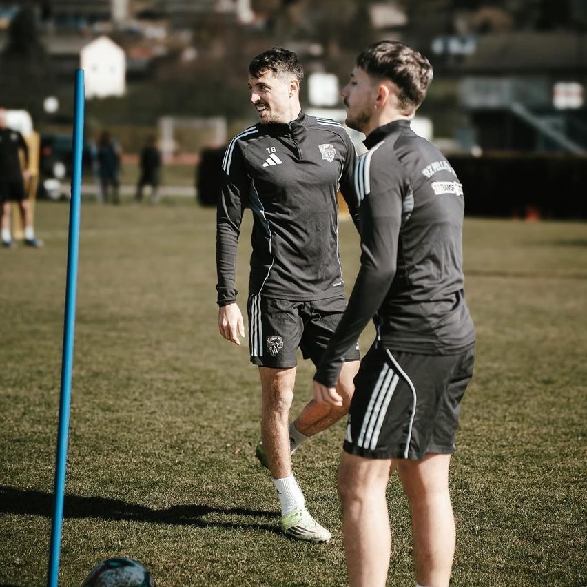 Two soccer players are conversing on the field. One wears a black Adidas jersey with a logo, while the other sports a long-sleeved Adidas shirt. A blue pole is nearby.
