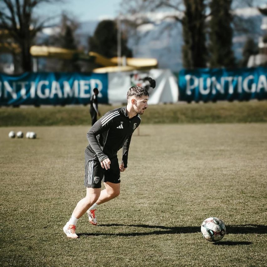 A soccer player practices on a field, with several soccer balls and a slide in the background. He wears a black jersey with white stripes.