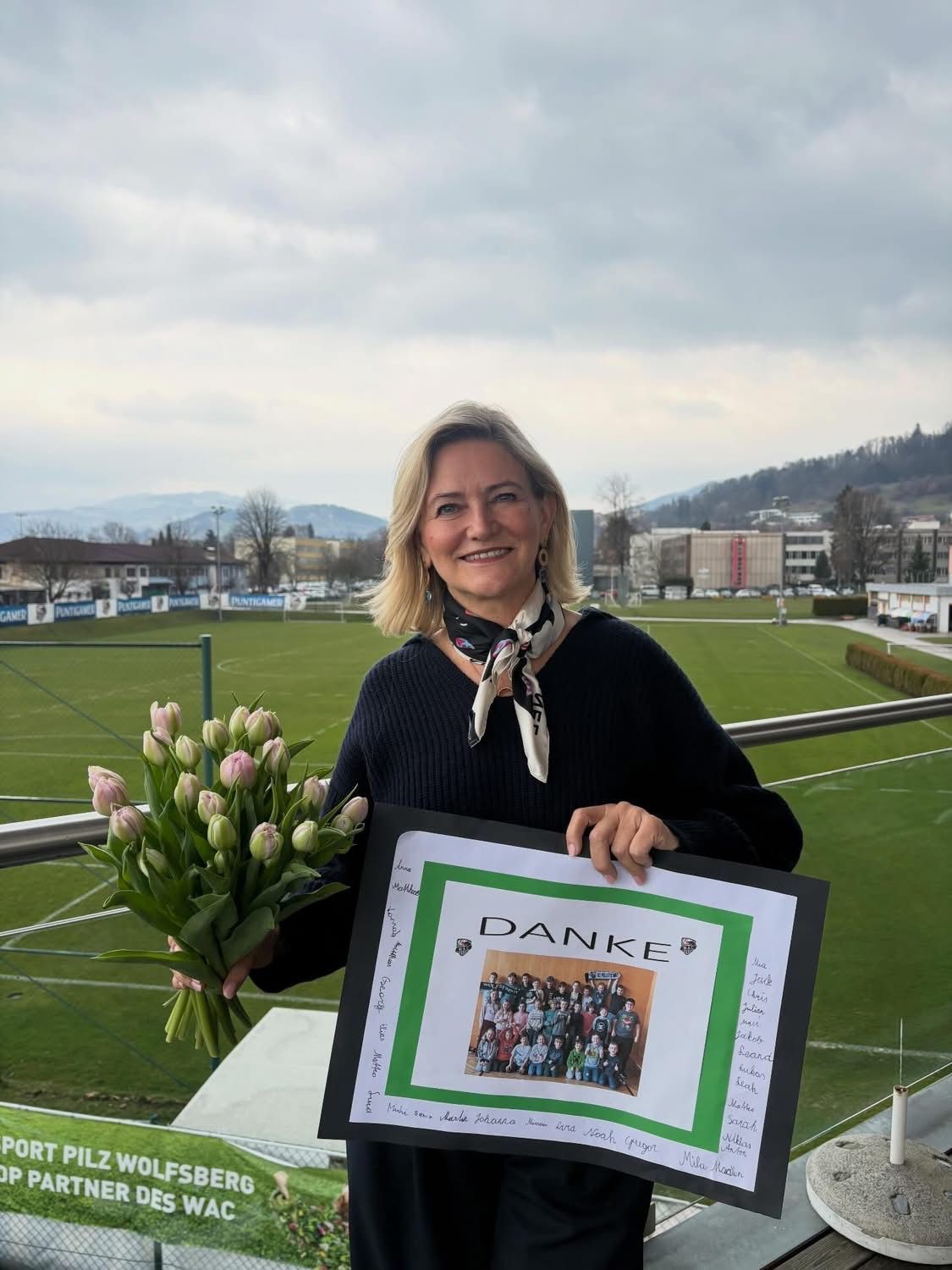 Eine Frau hält Blumen und ein Schild mit der Aufschrift Danke. Hinter ihr ist ein Gruppenfoto angebracht. Das Foto zeigt Menschen vor einem Feld mit einem Zaun.
