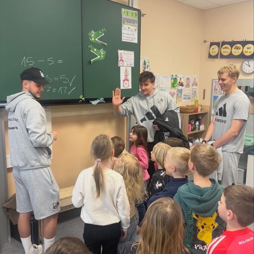 Drei Männer in grauen Trainingsanzüge stehen in einem Klassenzimmer mit Kindern. Ein Mann winkt, während ein anderer eine Jacke hält. Kinder versammeln sich und schauen zu den Erwachsenen auf. Eine Tafel zeigt Matheaufgaben an.