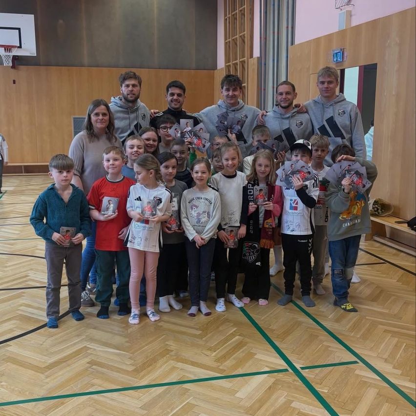 Eine Gruppe junger Kinder und Erwachsener posiert für ein Foto in einer Turnhalle, während sie signierte Gegenstände halten. Die Kinder sind in verschiedene Kleidungsstücke gekleidet, und die Erwachsenen tragen Kapuzenpullover. Ein Basketballkorb ist im Hintergrund zu sehen.