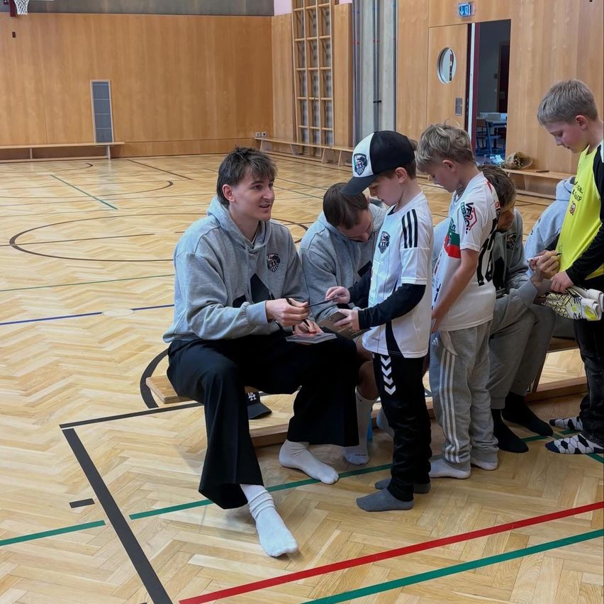 Ein Mann in einer Kapuzenjacke sitzt auf einer Bank in einer Turnhalle, hält ein Telefon und ist von mehreren Jungen umgeben. Die Jungen schauen auf das Telefon. Die Turnhalle hat Holzböden und Holzwände.