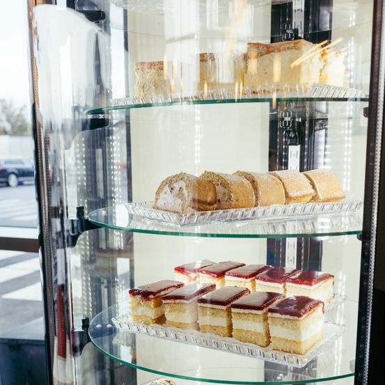 Eine Glasvitrine mit drei Ebenen zeigt eine Auswahl an Gebäck. Die oberste Ebene hat einen mehrschichtigen Kuchen. Die mittlere Ebene enthält eine Reihe von Sandwiches mit weißer Glasur. Die unterste Ebene zeigt ein Tablett mit kleinen, bunten Desserts.