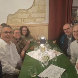 Eine Gruppe von Menschen sitzt um einen Tisch in einem Restaurant. Sie lächeln und schauen in die Kamera. Auf dem Tisch stehen Gläser, eine Speisekarte und eine Lampe. Hinter ihnen ist eine Ziegelsteinmauer.