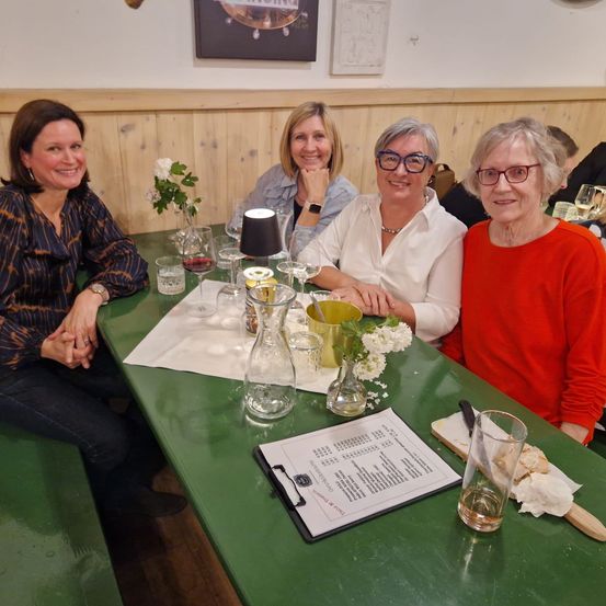 Vier Frauen sitzen an einem grünen Tisch und lächeln für ein Foto mit einem Menü vor ihnen. Der Tisch hat Weingläser, eine Vase mit Blumen und ein Notizbuch.