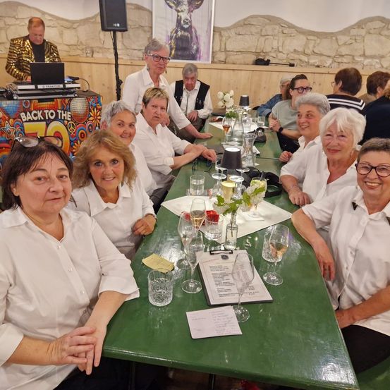 Eine Gruppe von Menschen sitzt an einem langen grünen Tisch mit Gläsern, einer Speisekarte und Blumen. Ein DJ spielt im Hintergrund.