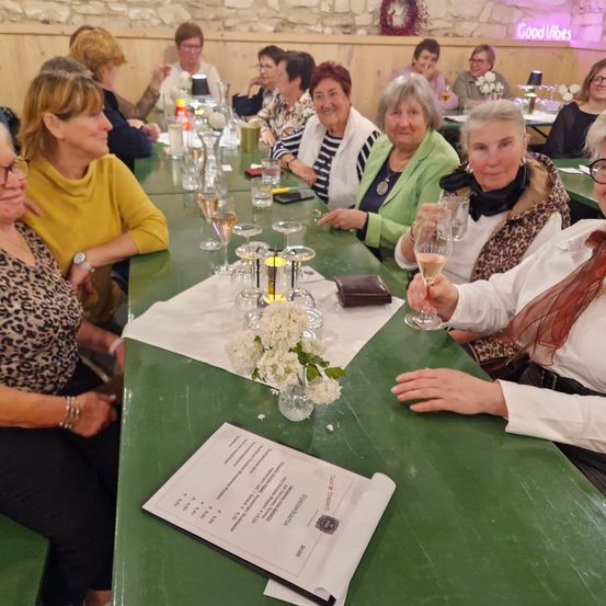 Eine Gruppe von Frauen sitzt um einen langen Tisch mit Blumen, Weingläsern und einer Speisekarte. Sie scheinen sich zu einem geselligen Beisammensein zu treffen.