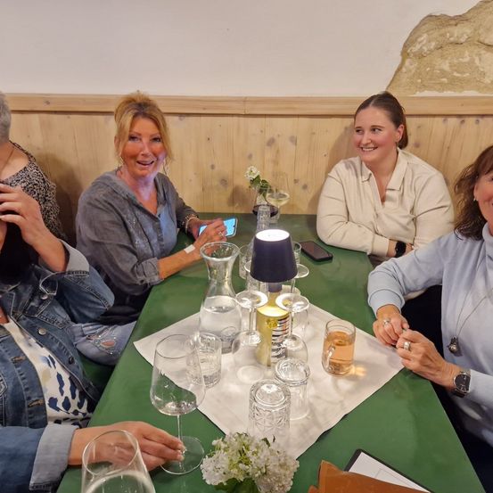 Vier Frauen sitzen um einen grünen Tisch und lächeln. Eine Frau benutzt ihr Handy, und eine Lampe steht auf dem Tisch. Es gibt Weingläser, eine Vase mit Blumen und eine Schüssel.